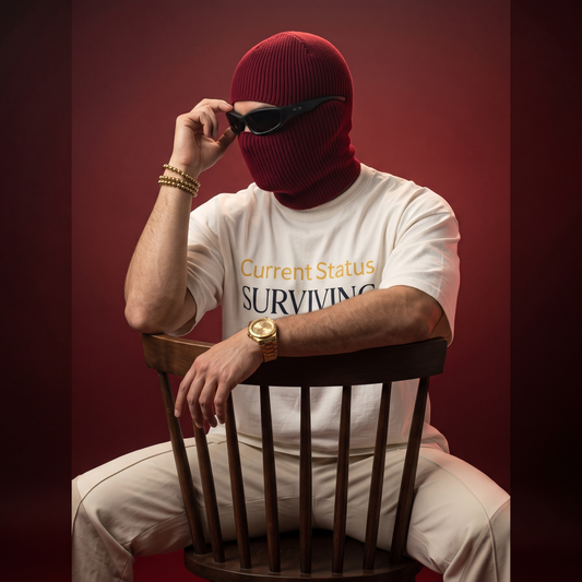 A person wearing a burgundy ribbed balaclava, black sunglasses, a white graphic T-shirt, and light beige pants sits on a dark wooden chair against a dark red studio background. The shirt reads 'Current Status SURVIVING'.