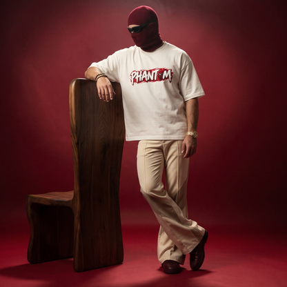 A person wearing a white graphic T-shirt with a red and black 'PHANTOM' logo, beige pants, and a burgundy balaclava leans against a wooden stool in a studio setting with a deep red background.