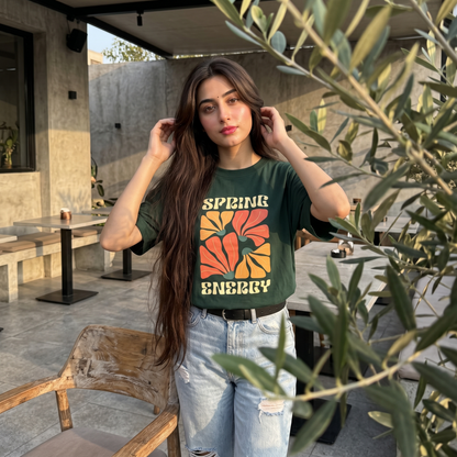 A woman posing outdoors in a dark green graphic t-shirt with a retro floral design that reads 'SPRING ENERGY' in yellow and orange, paired with light-wash distressed jeans.