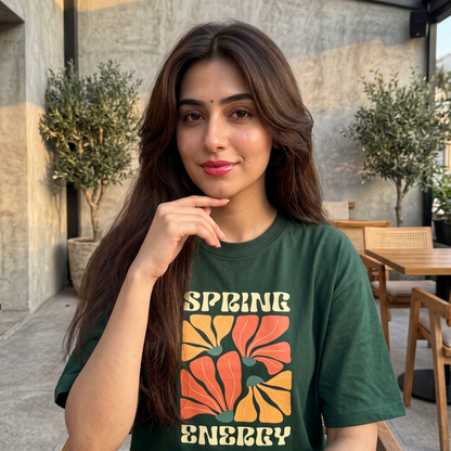A woman with long brown hair posing in a dark green graphic t-shirt that reads 'SPRING ENERGY' with a retro floral design in orange and yellow. She is standing in an outdoor patio setting with concrete walls and potted plants.