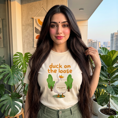 A woman with long dark hair stands on a balcony wearing a cream-colored graphic t-shirt with a cartoon duck and the text 'duck on the loose'. She is wearing olive green pants, with potted plants and a city skyline visible in the background.