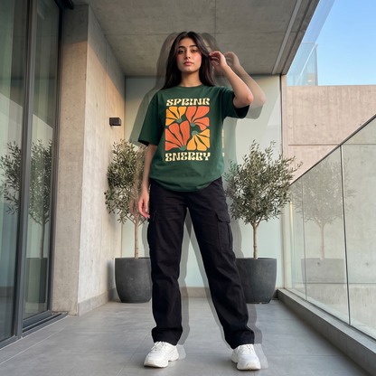A woman stands on a modern concrete balcony wearing a dark green graphic t-shirt with a retro orange and yellow sun design and the text 'SPINK ENJYY', paired with black cargo pants and white sneakers.