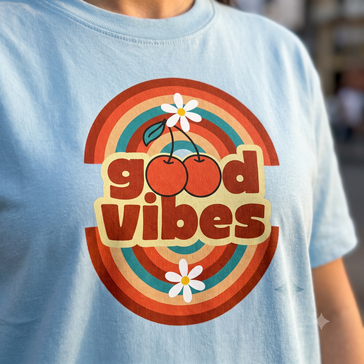 A close-up of a person wearing a light blue t-shirt with a retro 70s-style graphic that reads 'good vibes' in red and beige bubble letters, surrounded by orange, red, and teal concentric arches, with two cherries and white daisies.