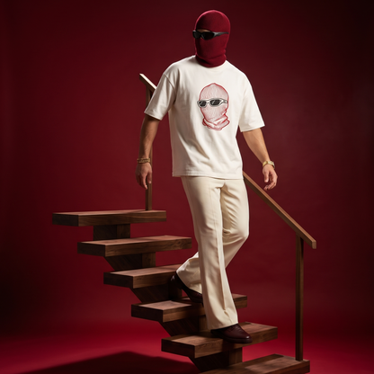 A person wearing a white graphic T-shirt, cream pants, and a burgundy balaclava stands on wooden stairs against a deep red studio background. The shirt has a stylized face graphic on the chest.