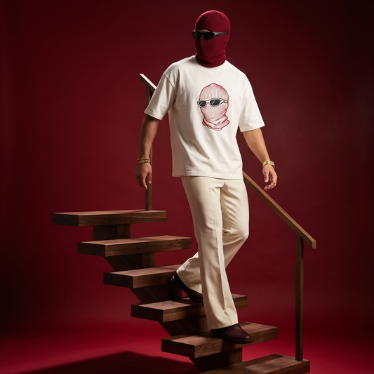 A person wearing a white graphic T-shirt, cream pants, and a burgundy balaclava stands on wooden stairs against a deep red studio background. The shirt has a stylized face graphic on the chest.