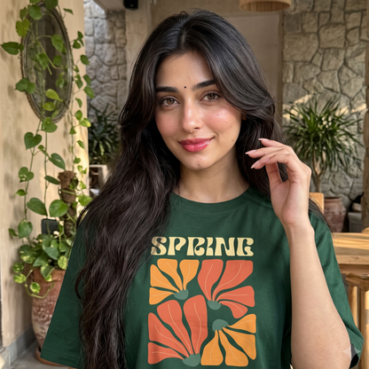 A woman with long dark hair wearing a dark green graphic t-shirt with a retro floral design and the words 'SPRING ENERGY' printed on the front, set in a rustic patio environment.