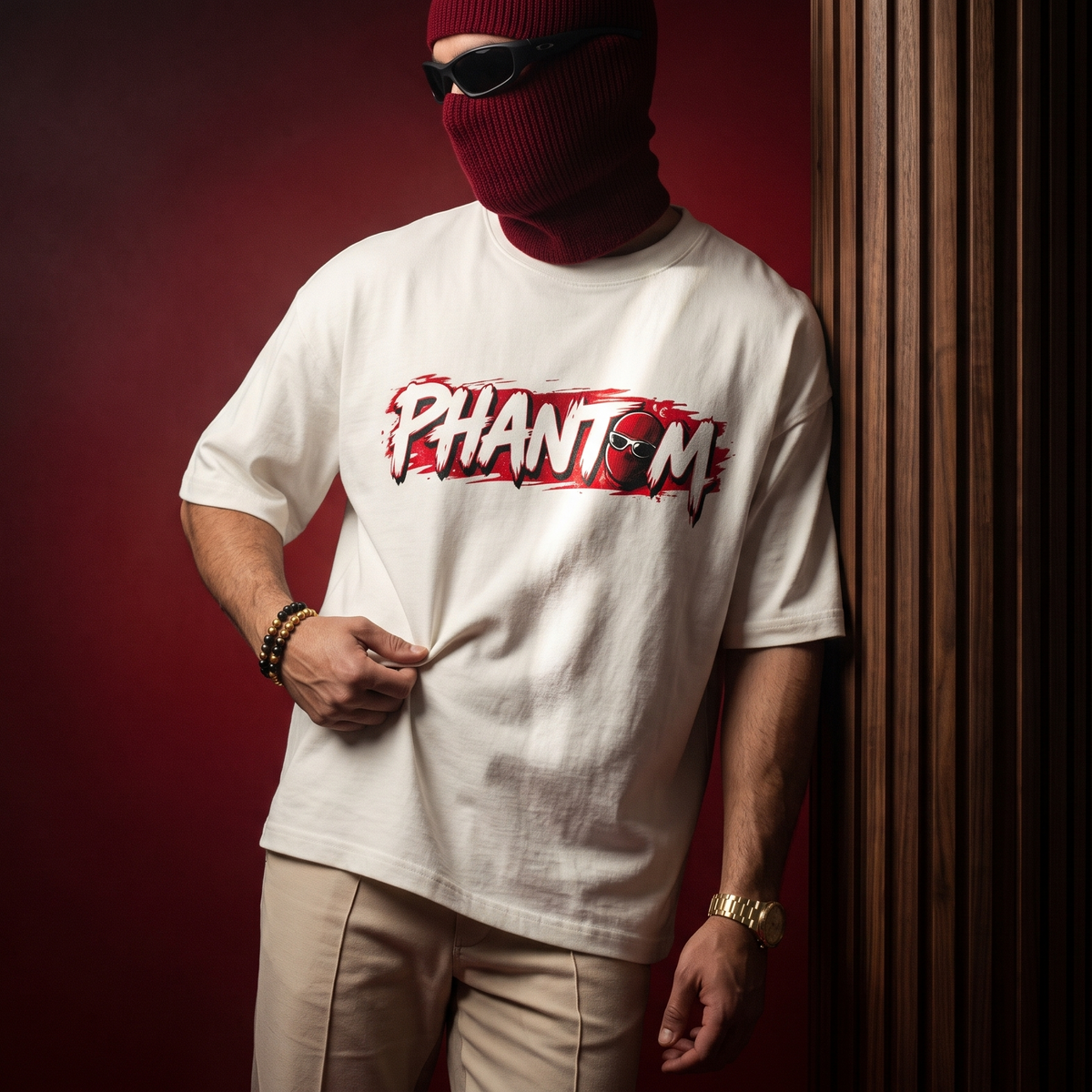 A person wearing a white graphic T-shirt with a bold red and black 'PHANTOM' logo across the chest, paired with beige pants and a dark red balaclava, standing against a dark red background.
