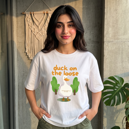A woman wearing a white graphic t-shirt with a cartoon duck and the text 'duck on the loose' printed on the front, standing in front of a concrete wall with a macrame wall hanging and a plant.