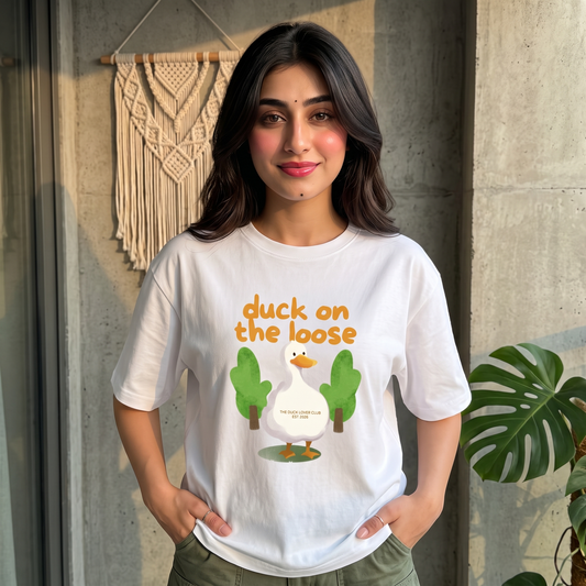 A woman wearing a white graphic t-shirt with a cartoon duck and the text 'duck on the loose' printed on the front, standing in front of a concrete wall with a macrame wall hanging and a plant.