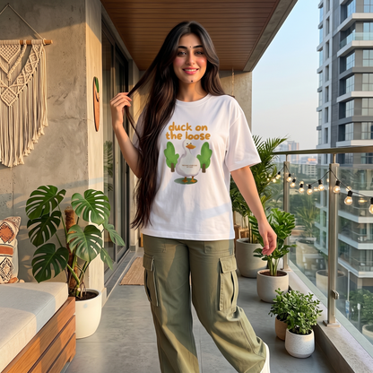A woman stands on a modern balcony wearing a white graphic T-shirt with the text 'duck on the loose' and a cactus illustration, paired with olive green cargo pants. The background features a contemporary high-rise building, potted plants, and warm string lights.