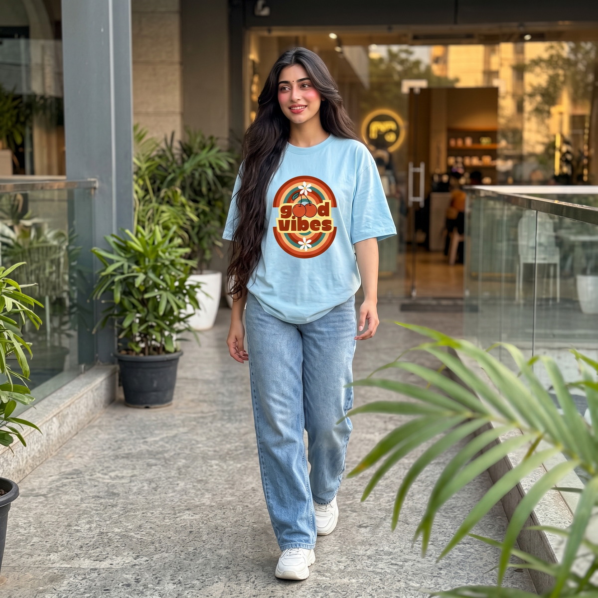 A woman walking in a modern indoor corridor wearing a light blue oversized graphic T-shirt with a circular retro-style logo that reads 'good vibes' and light-wash jeans.