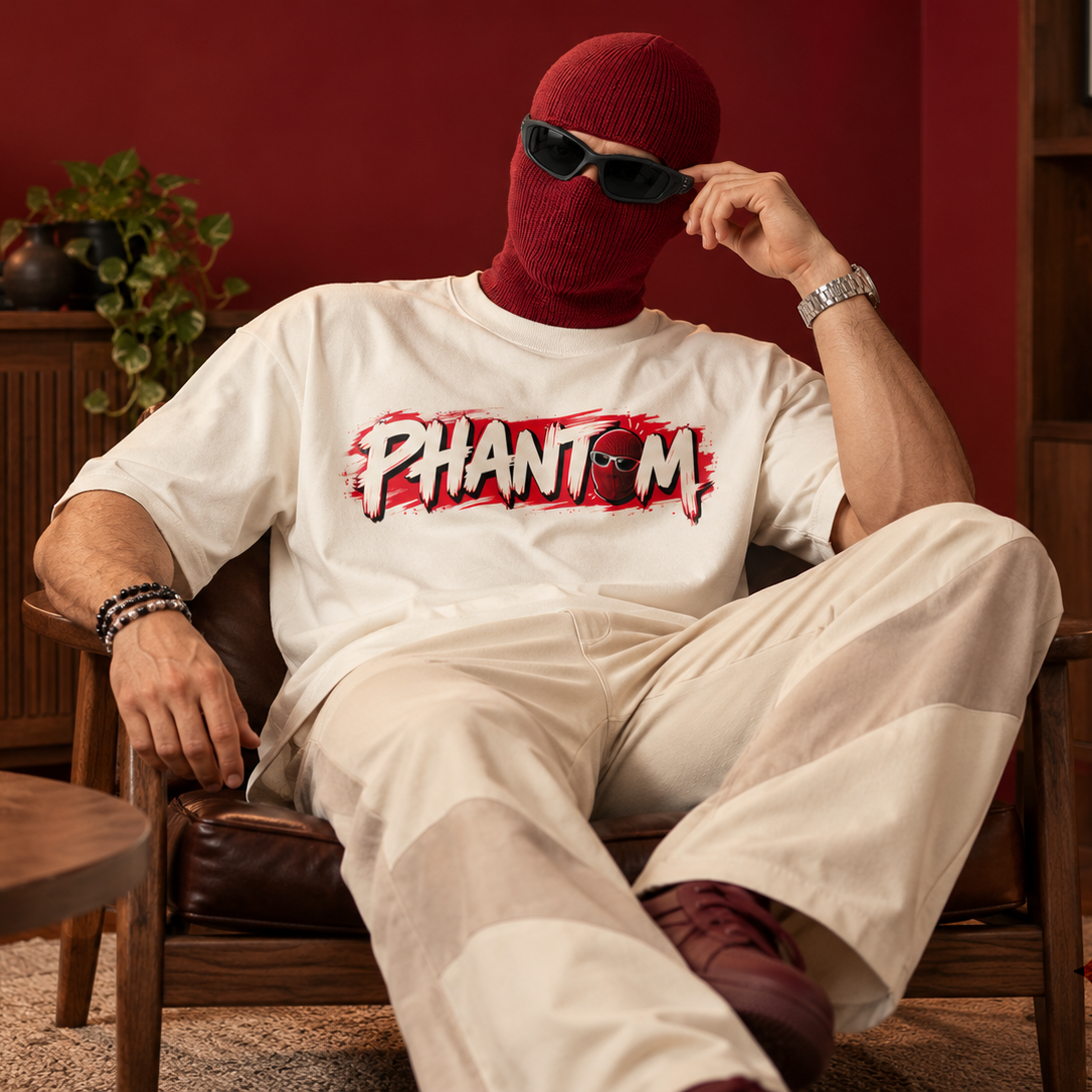 A person wearing a red ski mask and sunglasses sits in a brown leather armchair in a styled room with a red wall. They are wearing a white graphic T-shirt with 'PHANTOM' printed across the chest in red and black, paired with off-white pants.